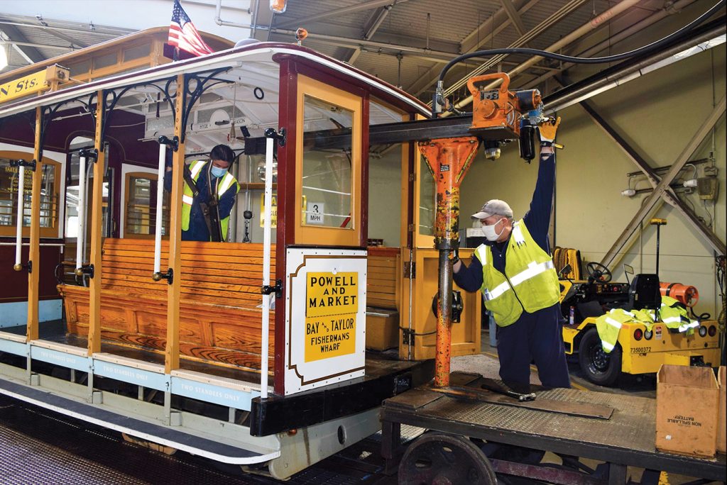 Two workers in high-visibility vests and masks are performing maintenance on a Powell and Market cable car inside a workshop. One worker is inside the cable car, working with tools, while the other is outside, using a large piece of equipment attached to the car. The cable car is stationed indoors, with an American flag on top and signs reading 'Powell and Market' and 'Bay & Taylor, Fisherman's Wharf.'
